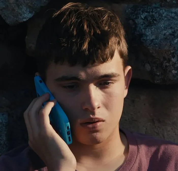 Eloy Pohu como Enzo no filme autointitulado, segurando um celular e preocupado sentado contra um muro de pedra.