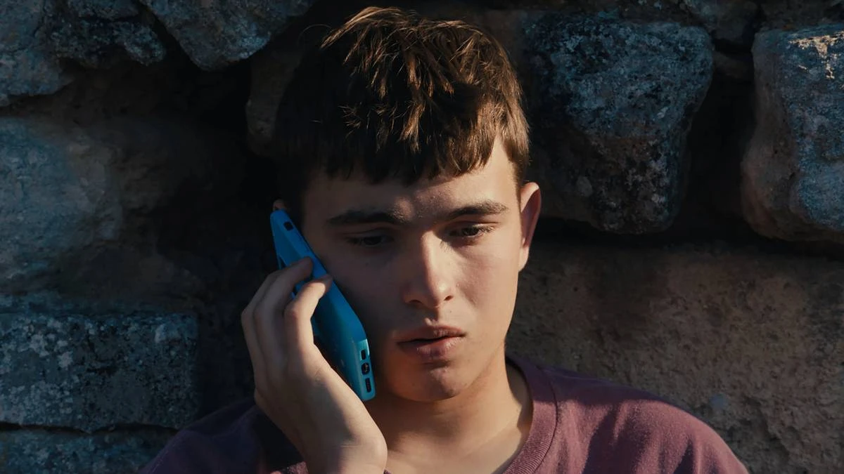 Eloy Pohu como Enzo no filme autointitulado, segurando um celular e preocupado sentado contra um muro de pedra.