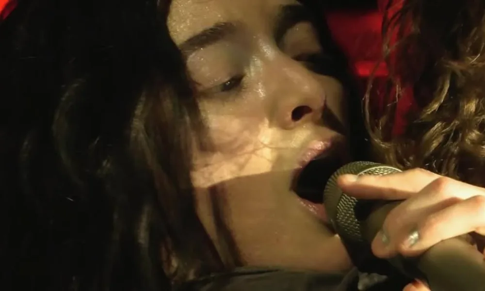 Lorde como vista por câmera capturando seu rosto com microfone bem próximo à boca em show, dando close na face.