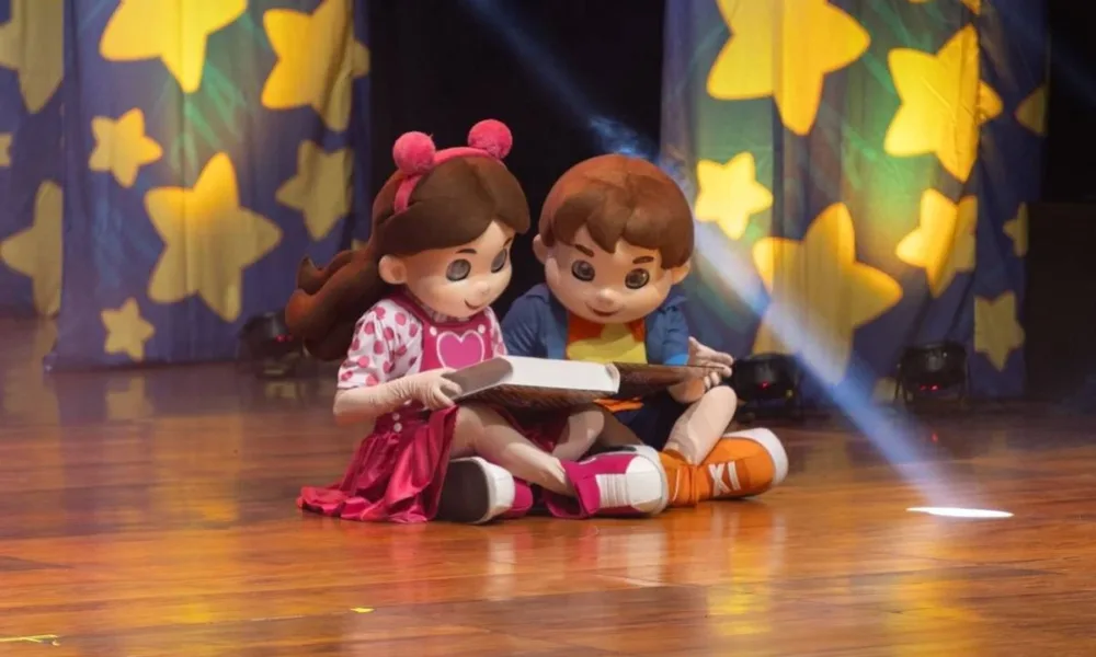 Bonecos Maria Clara & JP em espetáculo homônimo sentados no chão lendo um livro.