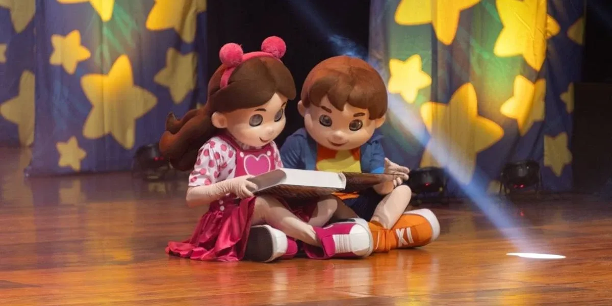 Bonecos Maria Clara & JP em espetáculo homônimo sentados no chão lendo um livro.