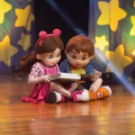 Bonecos Maria Clara & JP em espetáculo homônimo sentados no chão lendo um livro.