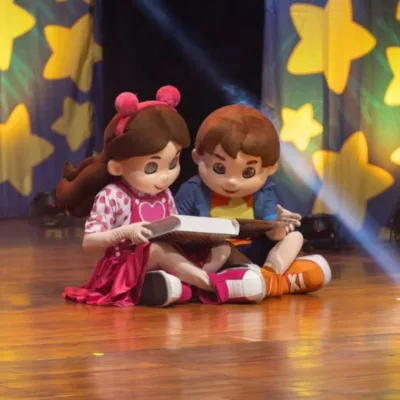 Bonecos Maria Clara & JP em espetáculo homônimo sentados no chão lendo um livro.