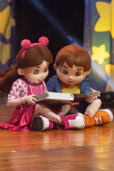 Bonecos Maria Clara & JP em espetáculo homônimo sentados no chão lendo um livro.