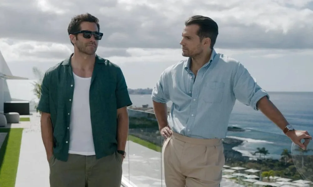 Jake Gyllenhaal (à esquerda) com mãos no bolso, de óculos escuros, e Henry Cavill (à direita) com mão na cintura e encostado olhando o ator ao lado, em paisagem de mansão no filme "Na Zona Cinzenta".