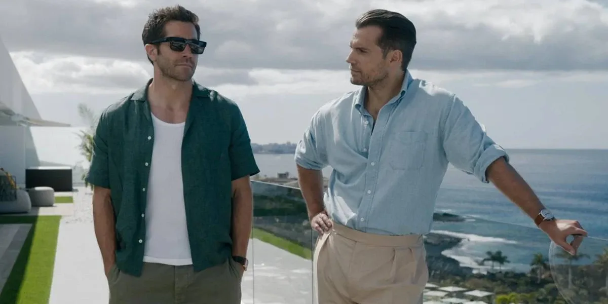 Jake Gyllenhaal (à esquerda) com mãos no bolso, de óculos escuros, e Henry Cavill (à direita) com mão na cintura e encostado olhando o ator ao lado, em paisagem de mansão no filme "Na Zona Cinzenta".