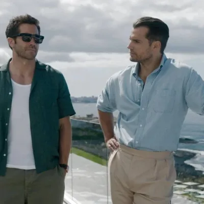 Jake Gyllenhaal (à esquerda) com mãos no bolso, de óculos escuros, e Henry Cavill (à direita) com mão na cintura e encostado olhando o ator ao lado, em paisagem de mansão no filme "Na Zona Cinzenta".