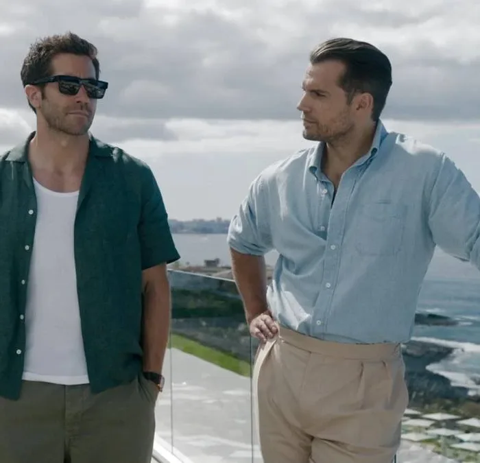 Jake Gyllenhaal (à esquerda) com mãos no bolso, de óculos escuros, e Henry Cavill (à direita) com mão na cintura e encostado olhando o ator ao lado, em paisagem de mansão no filme "Na Zona Cinzenta".