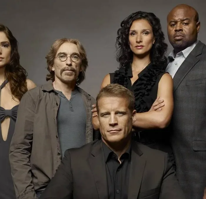 Elenco de "Screener: Alvo Humano" com protagonista ao centor centado, enquanto o resto do elenco está em pé posando para foto promocional em fundo cinza caracterizados como seus personagens na série.