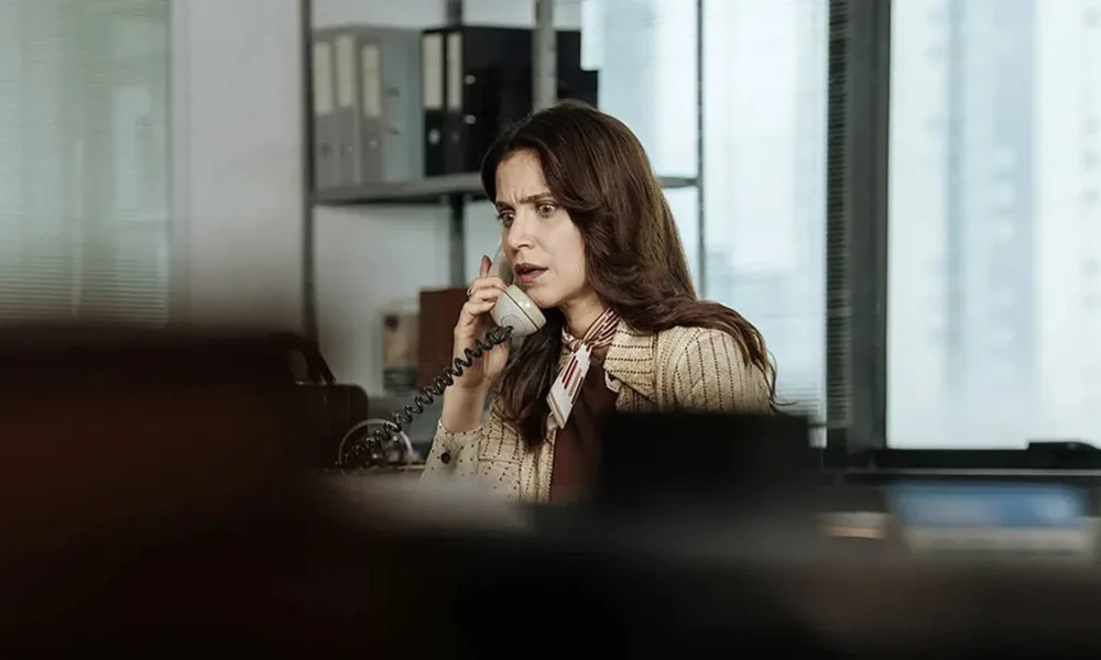 Mel Lisboa como a protagonista de "A Conspiração Condor" em escritório, atendendo telefone, com expressão de susto; câmera aponta à personagem da atriz por detrás de uma pilha de objetos, dando profundidade de estar sendo espionada.