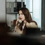 Mel Lisboa como a protagonista de "A Conspiração Condor" em escritório, atendendo telefone, com expressão de susto; câmera aponta à personagem da atriz por detrás de uma pilha de objetos, dando profundidade de estar sendo espionada.