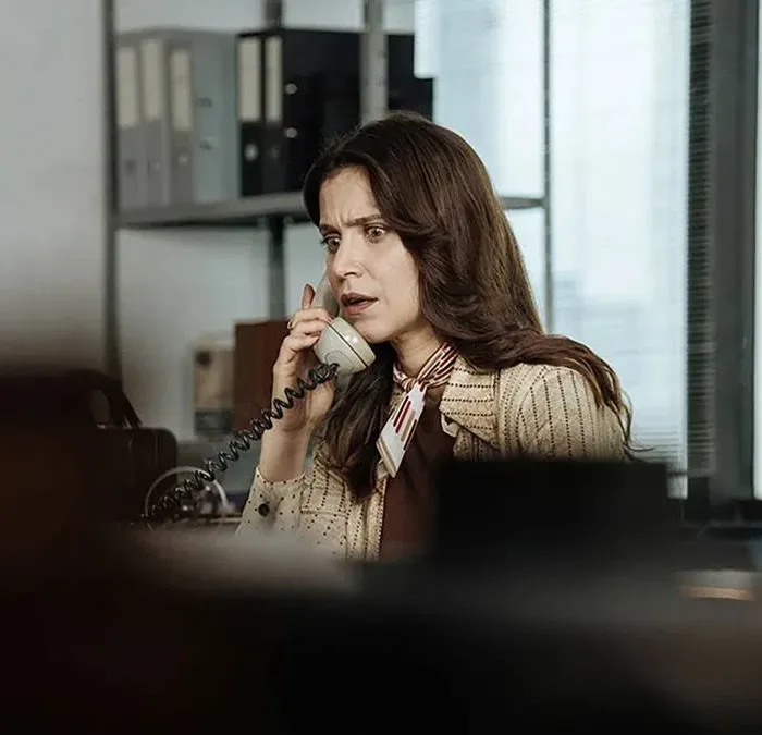 Mel Lisboa como a protagonista de "A Conspiração Condor" em escritório, atendendo telefone, com expressão de susto; câmera aponta à personagem da atriz por detrás de uma pilha de objetos, dando profundidade de estar sendo espionada.