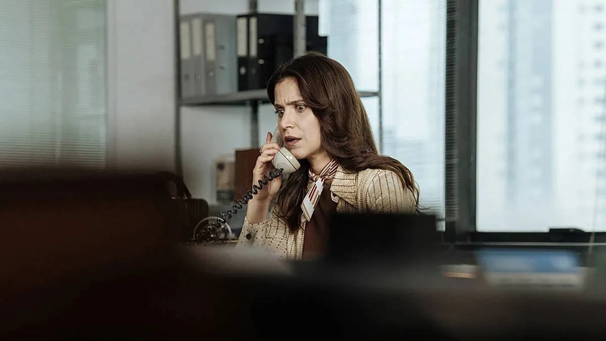 A Conspiração Condor | Até Quando Precisaremos de Filmes Sobre a Ditadura? 2 Mel Lisboa como a protagonista de "A Conspiração Condor" em escritório, atendendo telefone, com expressão de susto; câmera aponta à personagem da atriz por detrás de uma pilha de objetos, dando profundidade de estar sendo espionada.