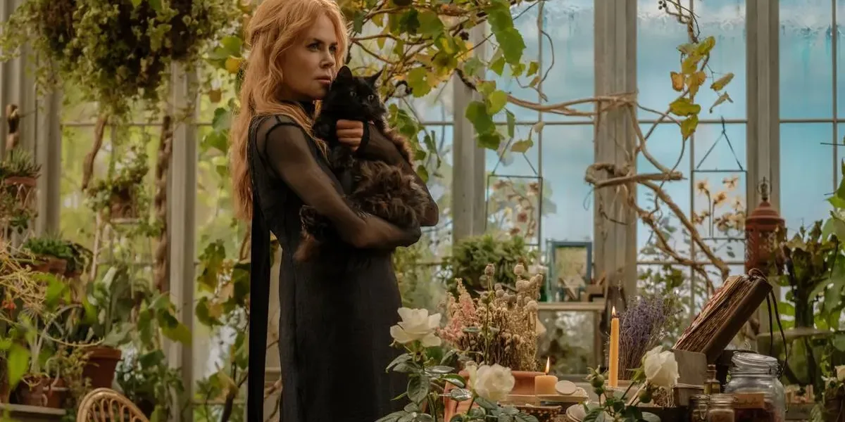 Nicole Kidman como a bruxa Gillian Owens na sequência "Da Magia à Sedução 2". Personagem está com cara fechada, segurando um gato preto no colo com os dois braços, em pé, vista de lado. Ambientação de estufa caseira cheia de plantas.