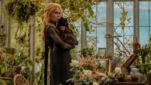 Nicole Kidman como a bruxa Gillian Owens na sequência "Da Magia à Sedução 2". Personagem está com cara fechada, segurando um gato preto no colo com os dois braços, em pé, vista de lado. Ambientação de estufa caseira cheia de plantas.