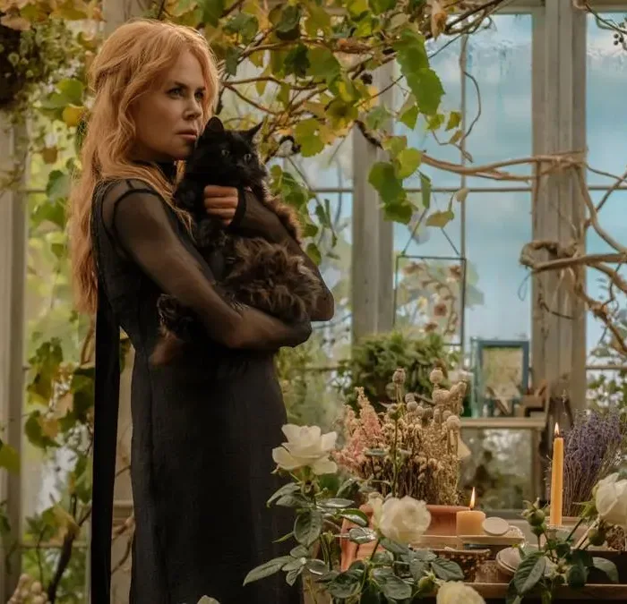 Nicole Kidman como a bruxa Gillian Owens na sequência "Da Magia à Sedução 2". Personagem está com cara fechada, segurando um gato preto no colo com os dois braços, em pé, vista de lado. Ambientação de estufa caseira cheia de plantas.