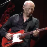 Mark Knopfler durante apresentação da Shangri-La Tour em 2005. Músico está vestido de preto e olhando para sua guitarra.