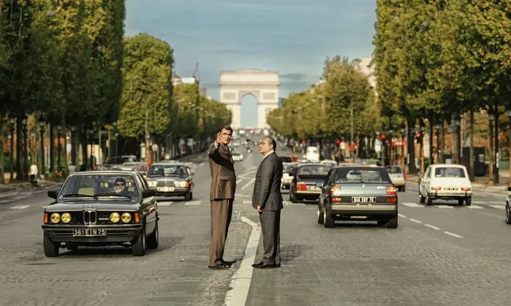 Foto horizontal promocional para o longa "O Grande Arco de Paris", com protagonistas no meio de avenida com Arco do Triunfo atrás. Imagem também é a mesma do pôster.