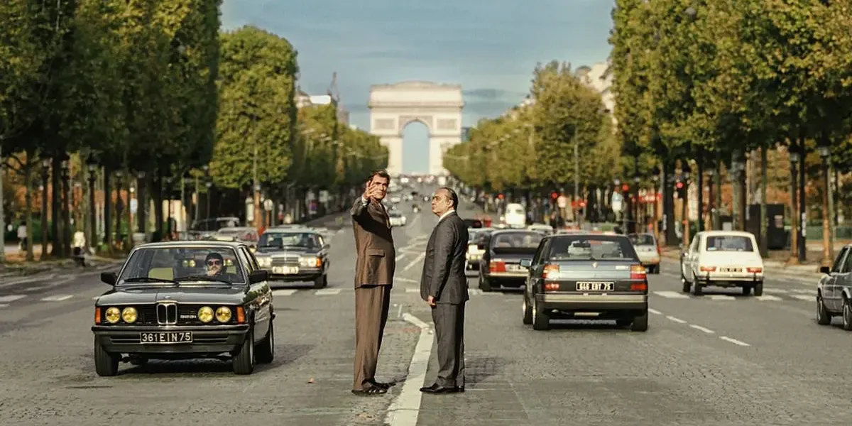 Foto horizontal promocional para o longa "O Grande Arco de Paris", com protagonistas no meio de avenida com Arco do Triunfo atrás. Imagem também é a mesma do pôster.