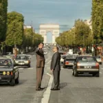 Foto horizontal promocional para o longa "O Grande Arco de Paris", com protagonistas no meio de avenida com Arco do Triunfo atrás. Imagem também é a mesma do pôster.