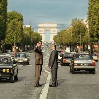 Foto horizontal promocional para o longa "O Grande Arco de Paris", com protagonistas no meio de avenida com Arco do Triunfo atrás. Imagem também é a mesma do pôster.