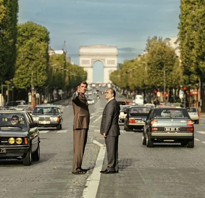 Foto horizontal promocional para o longa "O Grande Arco de Paris", com protagonistas no meio de avenida com Arco do Triunfo atrás. Imagem também é a mesma do pôster.