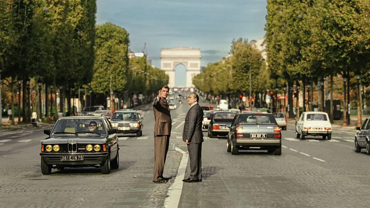 Foto horizontal promocional para o longa "O Grande Arco de Paris", com protagonistas no meio de avenida com Arco do Triunfo atrás. Imagem também é a mesma do pôster.
