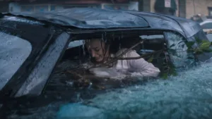 Phoebe Dynevor como Lisa Fields em "Ataque Brutal". Personagem está ensopada dentro de carro, um galho enorme entrando dentro do carro, e ela desesperada.