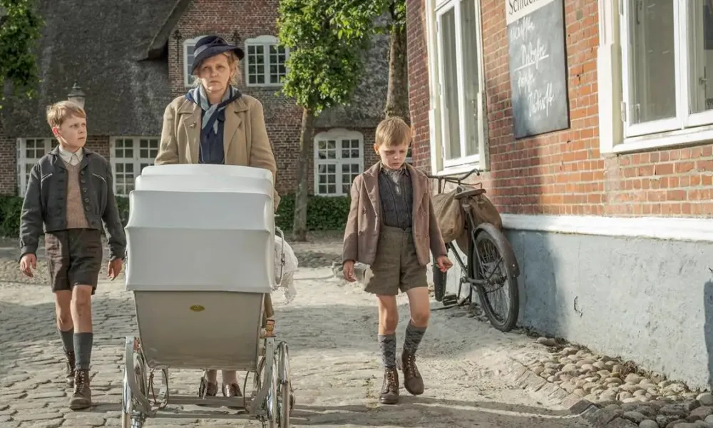 Cena do filme "Uma Infância Alemã". Mãe caminha com dois filhos, um a cada lado, mais o carrinho de bebê que puxa. Ela está com um semblante irritadiço e os garotos cabisbaixos.