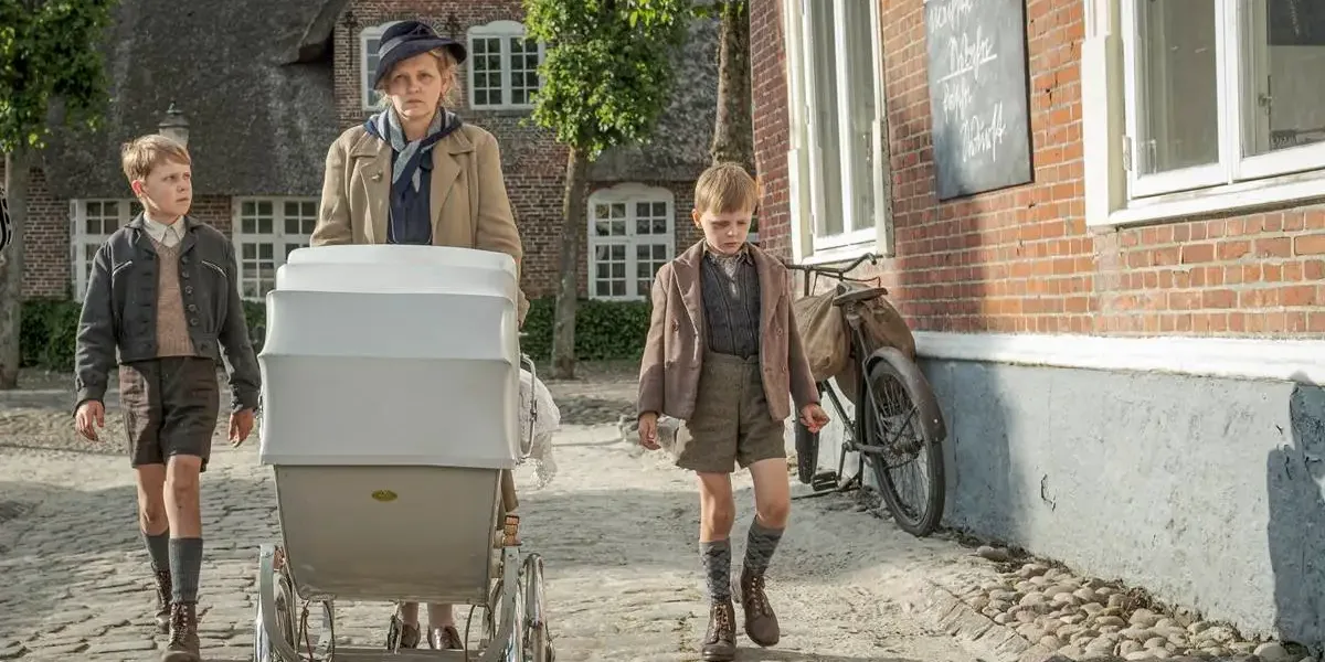Cena do filme "Uma Infância Alemã". Mãe caminha com dois filhos, um a cada lado, mais o carrinho de bebê que puxa. Ela está com um semblante irritadiço e os garotos cabisbaixos.