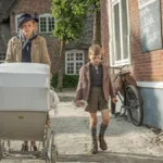 Cena do filme "Uma Infância Alemã". Mãe caminha com dois filhos, um a cada lado, mais o carrinho de bebê que puxa. Ela está com um semblante irritadiço e os garotos cabisbaixos.