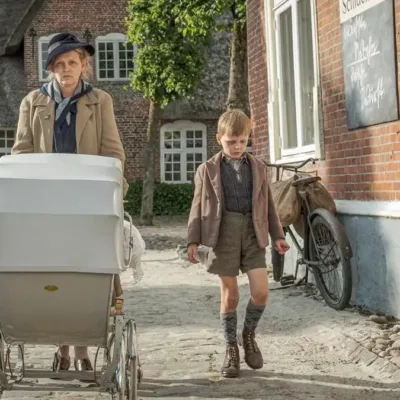 Cena do filme "Uma Infância Alemã". Mãe caminha com dois filhos, um a cada lado, mais o carrinho de bebê que puxa. Ela está com um semblante irritadiço e os garotos cabisbaixos.