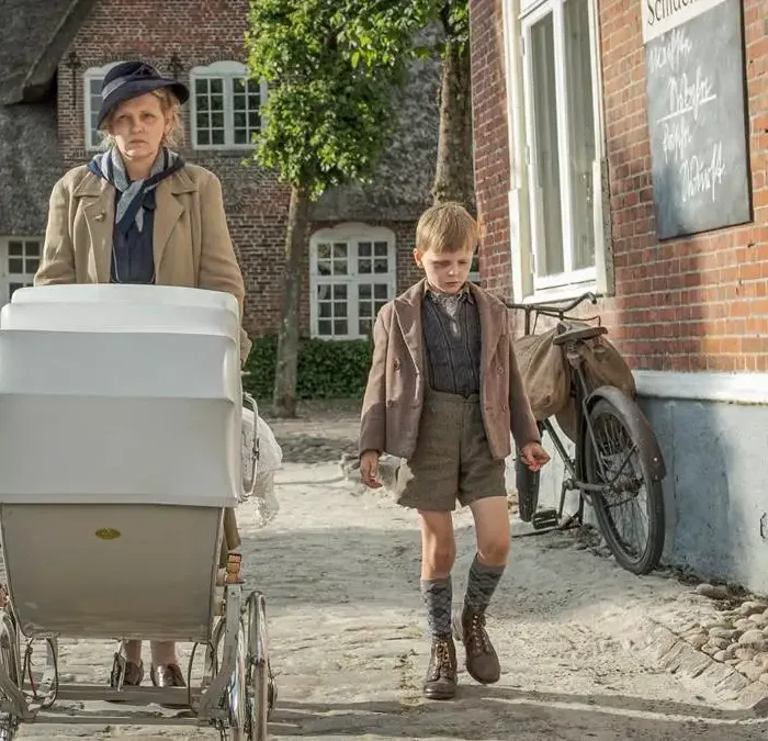 Cena do filme "Uma Infância Alemã". Mãe caminha com dois filhos, um a cada lado, mais o carrinho de bebê que puxa. Ela está com um semblante irritadiço e os garotos cabisbaixos.