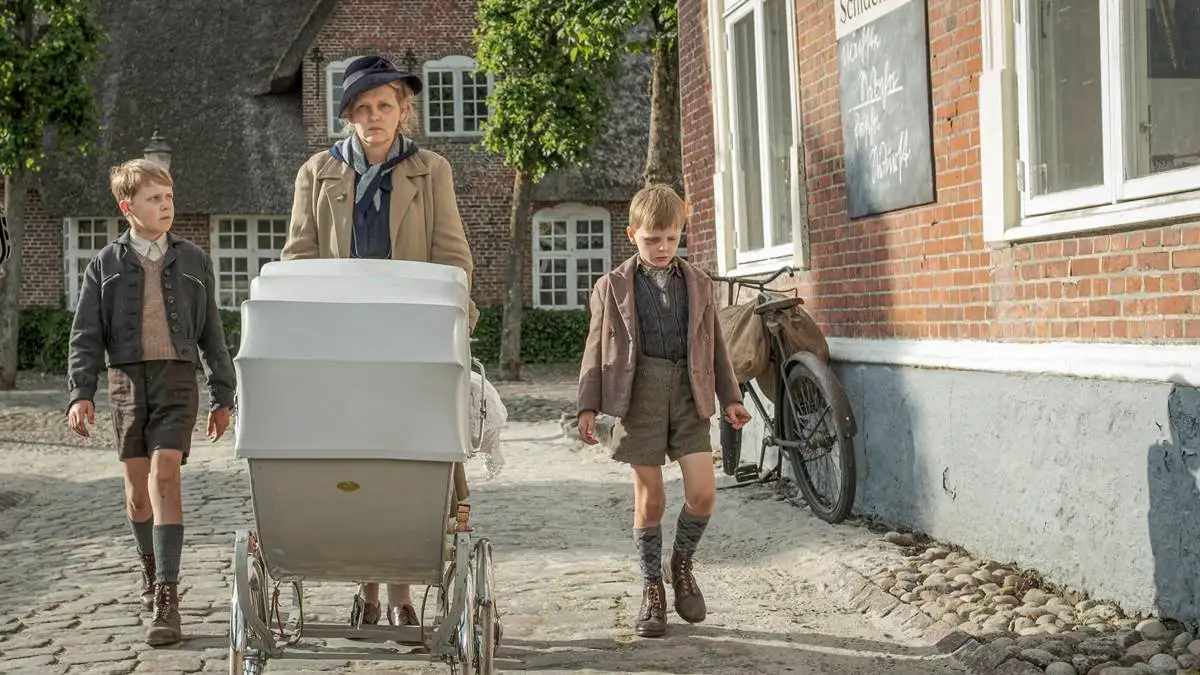 Cena do filme "Uma Infância Alemã". Mãe caminha com dois filhos, um a cada lado, mais o carrinho de bebê que puxa. Ela está com um semblante irritadiço e os garotos cabisbaixos.
