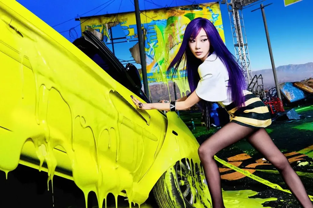 Esta imagem mostra Giselle, do grupo de K-pop aespa, em um ensaio fotográfico para o segundo álbum completo do grupo, intitulado "LEMONADE", inclinada passando a mão em um carro coberto de slime neon amarelo, os cabelos roxos da cantora também igualmente brilhantes.