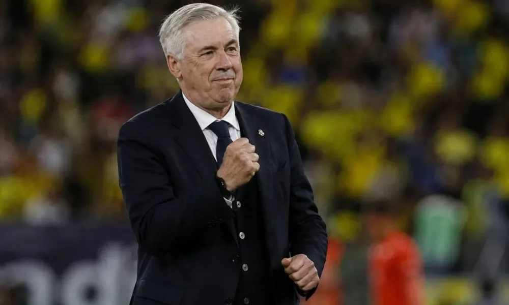 Carlo Ancelotti, técnico da seleção brasileira, punho esquerdo fechado e expressão em sinal de comemoração, terno e gravata em campo durante partida. Imagem gerada por IA.
