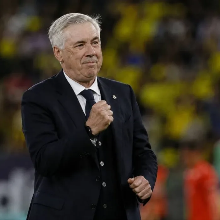 Carlo Ancelotti, técnico da seleção brasileira, punho esquerdo fechado e expressão em sinal de comemoração, terno e gravata em campo durante partida. Imagem gerada por IA.