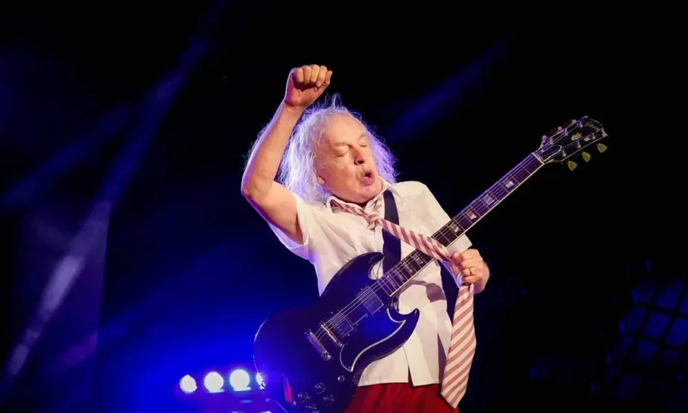 Angus Young, guitarrista do AC/DC, durante show em julho de 2024 em Nürnberg. Ele está com uma gravata e roupas escolares, braço direito levantado, pegando na gravata com a mão esquerda, durante performance.