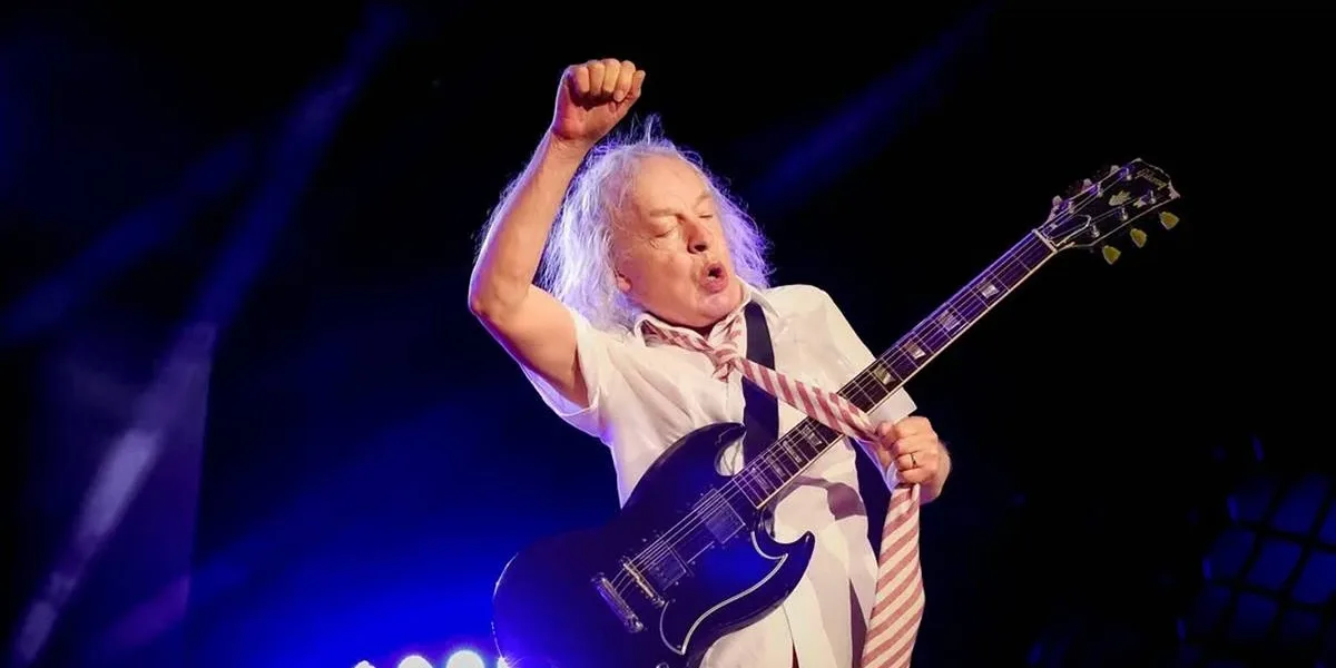 Angus Young, guitarrista do AC/DC, durante show em julho de 2024 em Nürnberg. Ele está com uma gravata e roupas escolares, braço direito levantado, pegando na gravata com a mão esquerda, durante performance.