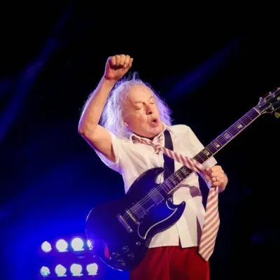 Angus Young, guitarrista do AC/DC, durante show em julho de 2024 em Nürnberg. Ele está com uma gravata e roupas escolares, braço direito levantado, pegando na gravata com a mão esquerda, durante performance.