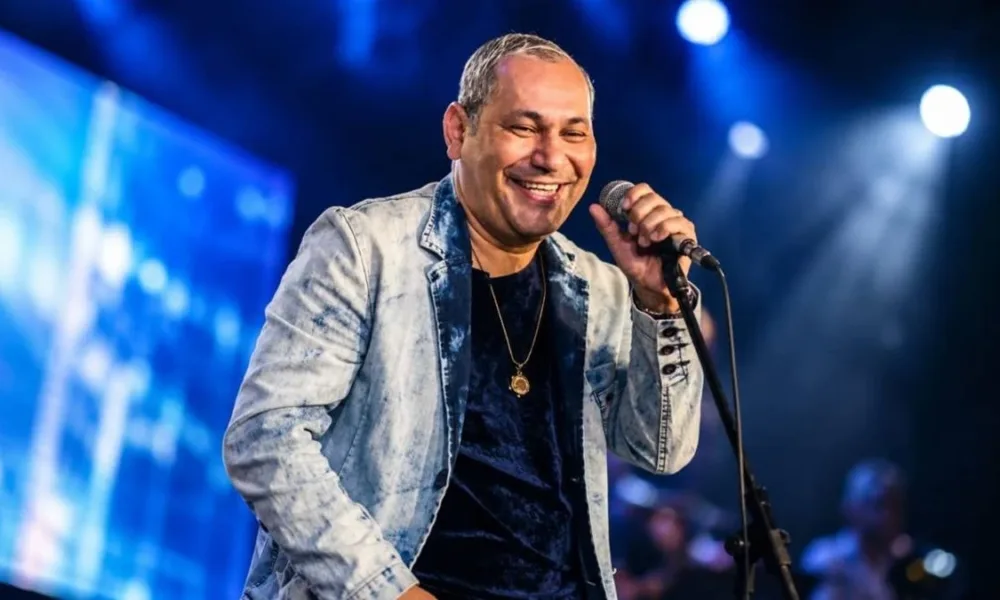 Cantor Chrigot durante apresentação, mão esquerda no microfone fixo, sorrindo, fundo com luzes e telão azul desfocado. Imagem gerada por IA.