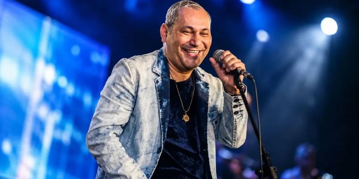 Chrigor é Internado Novamente e Cancela Apresentações no Sul do país Cantor Chrigot durante apresentação, mão esquerda no microfone fixo, sorrindo, fundo com luzes e telão azul desfocado. Imagem gerada por IA.