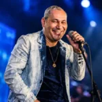 Cantor Chrigot durante apresentação, mão esquerda no microfone fixo, sorrindo, fundo com luzes e telão azul desfocado. Imagem gerada por IA.