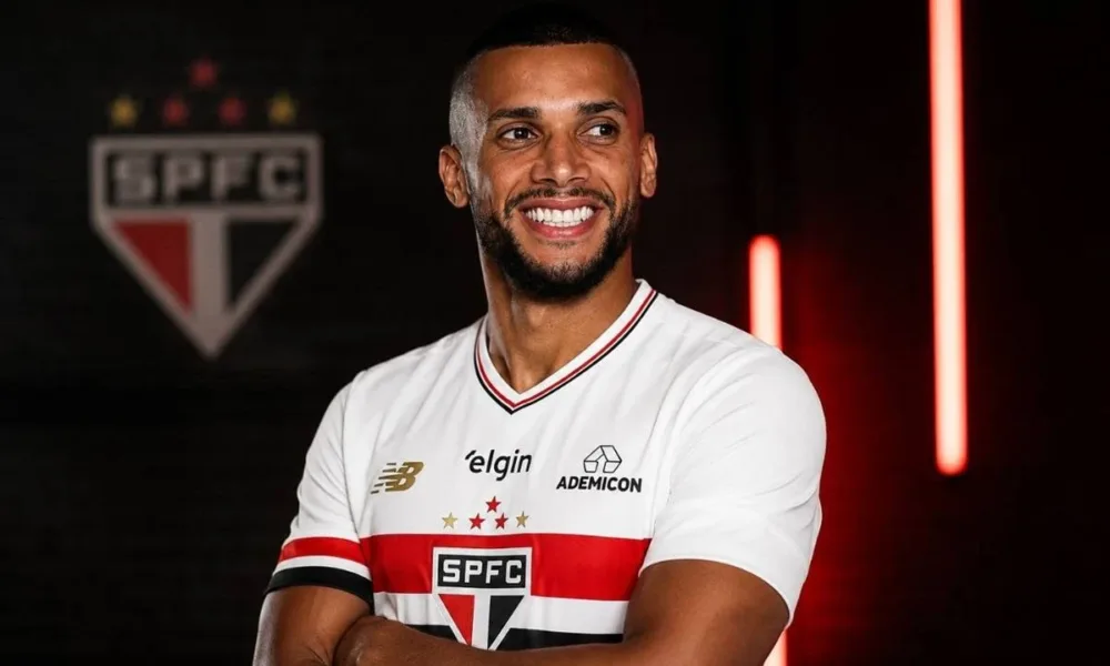 Dória, ex-jogador do São Paulo, com camisa do time, braços cruzados, em fundo escuro com iluminação morna vermelha e atrás o escudo do São Paulo. Ele sorri olhando distante.