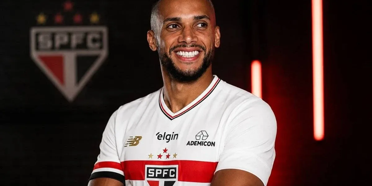 Ameaças, Pressão e Desgaste: a Crise que Levou Dória a Deixar o São Paulo Poucos Meses após Retorno Dória, ex-jogador do São Paulo, com camisa do time, braços cruzados, em fundo escuro com iluminação morna vermelha e atrás o escudo do São Paulo. Ele sorri olhando distante.