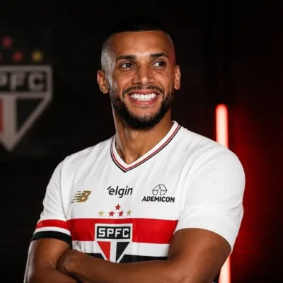 Dória, ex-jogador do São Paulo, com camisa do time, braços cruzados, em fundo escuro com iluminação morna vermelha e atrás o escudo do São Paulo. Ele sorri olhando distante.