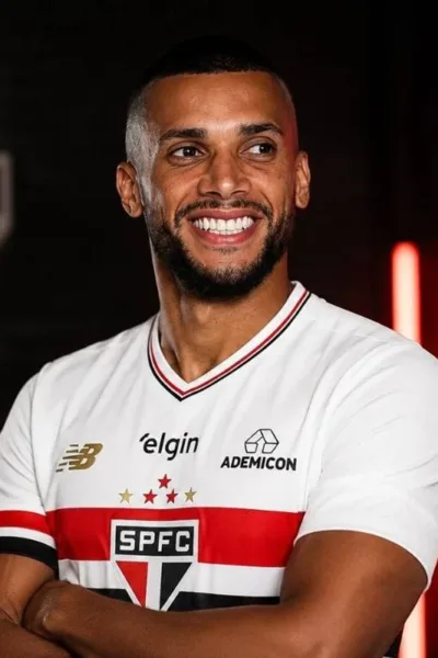 Dória, ex-jogador do São Paulo, com camisa do time, braços cruzados, em fundo escuro com iluminação morna vermelha e atrás o escudo do São Paulo. Ele sorri olhando distante.