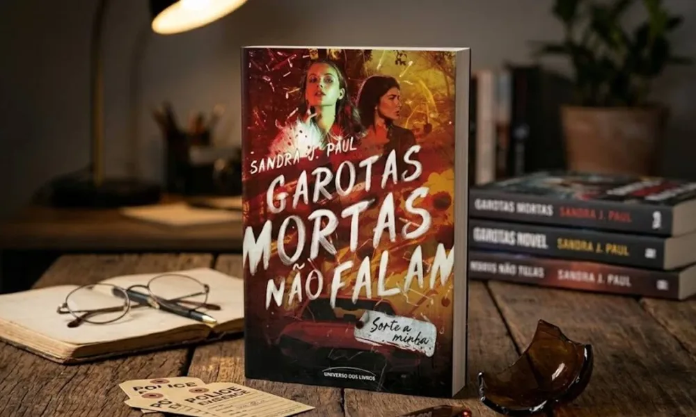 Livro "Garotas Mortas Não Falam" de Sandra J. Paul em destaque de pé em meio a mesa de madeira, livros outros da autora são vistos ao fundo. Há um caco de vidro em destaque também. Imagem gerada por IA.
