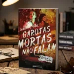 Livro "Garotas Mortas Não Falam" de Sandra J. Paul em destaque de pé em meio a mesa de madeira, livros outros da autora são vistos ao fundo. Há um caco de vidro em destaque também. Imagem gerada por IA.