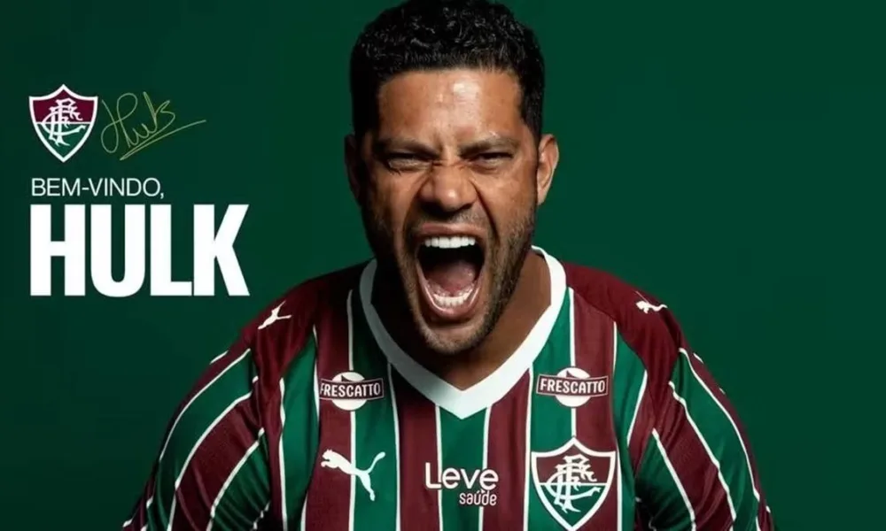 Atacante Hulk em imagem, horizontal, de anúncio de sua vinda para o Fluminense a partir de 2026. Ele está com a camisa do time e dá um grito. Escudo do time está acima de seu nome, à esquerda do fundo verde, gravado em branco.